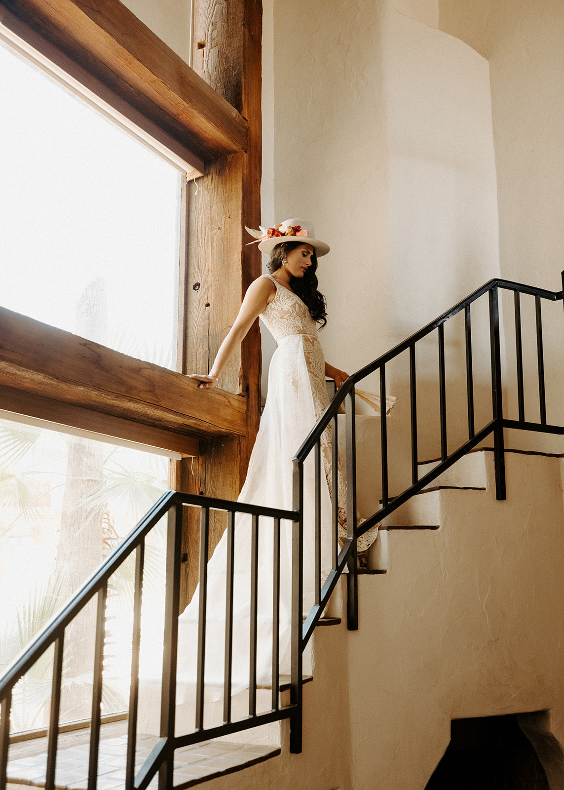 Bride in a wedding dress standing on a staircase with large windows in the background wearing a custom western hat.