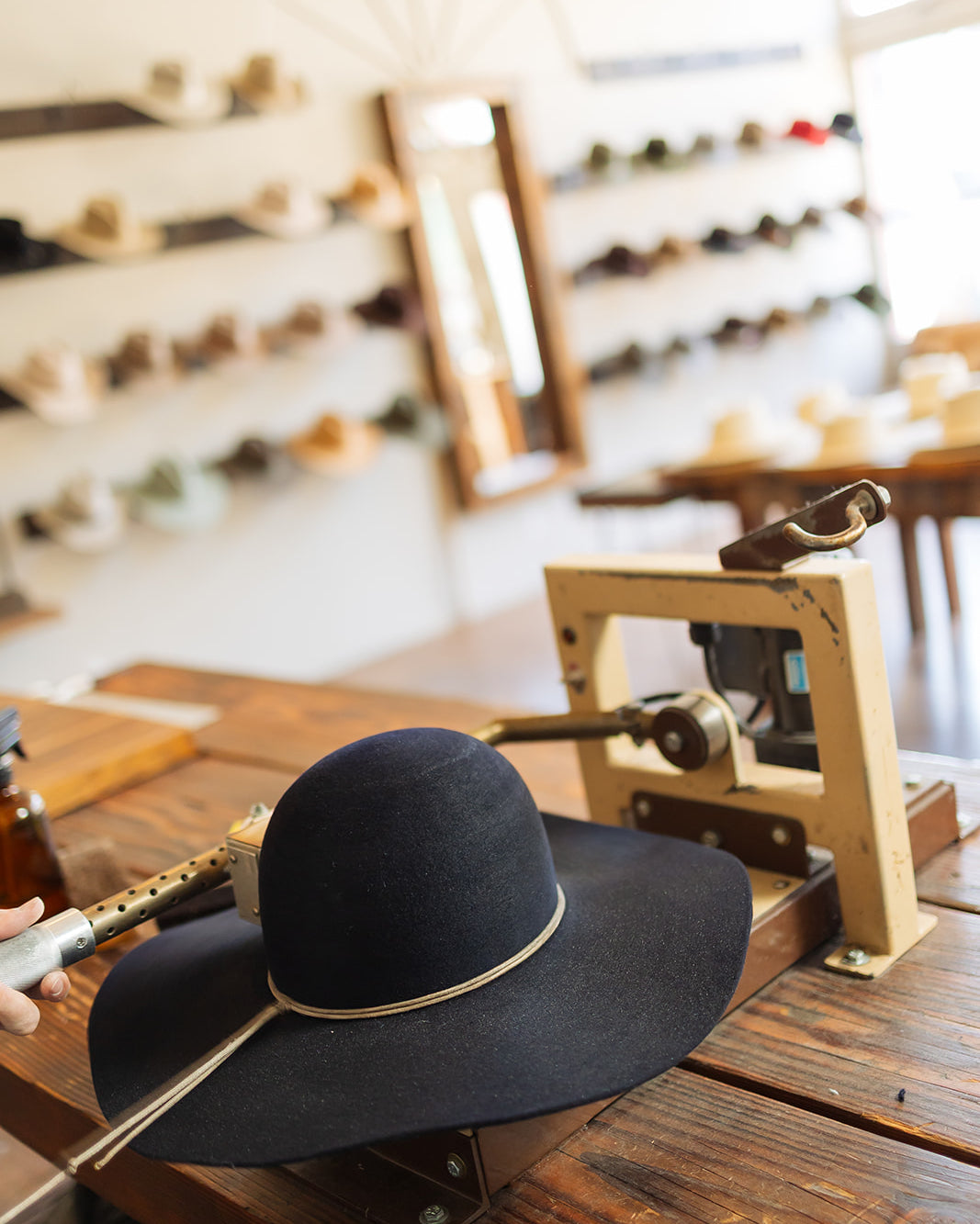 Black wide-brimmed hat with gold band on a wooden table in a workshop setting.