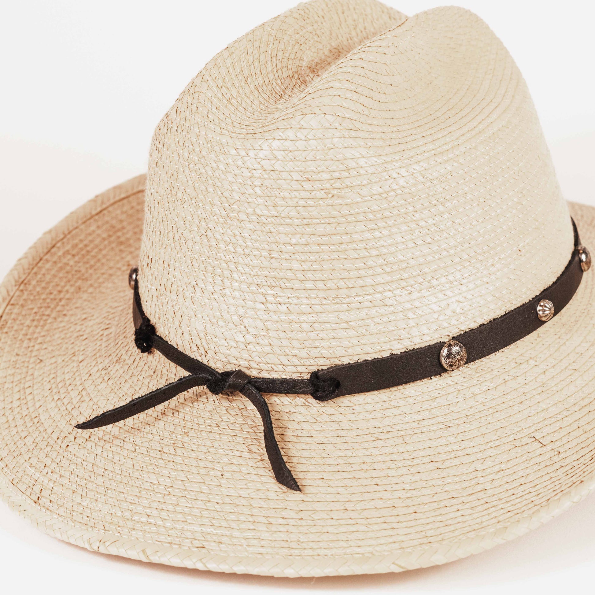 A light beige woven straw fedora hat with a dark brown ribbon band decorated with small metallic studs and tied in a bow at the front, shown on a plain white background.