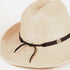A close-up of a woven straw western hat in a light beige color, featuring a black ribbon band tied in a bow and adorned with a small round silver-tone decorative button.