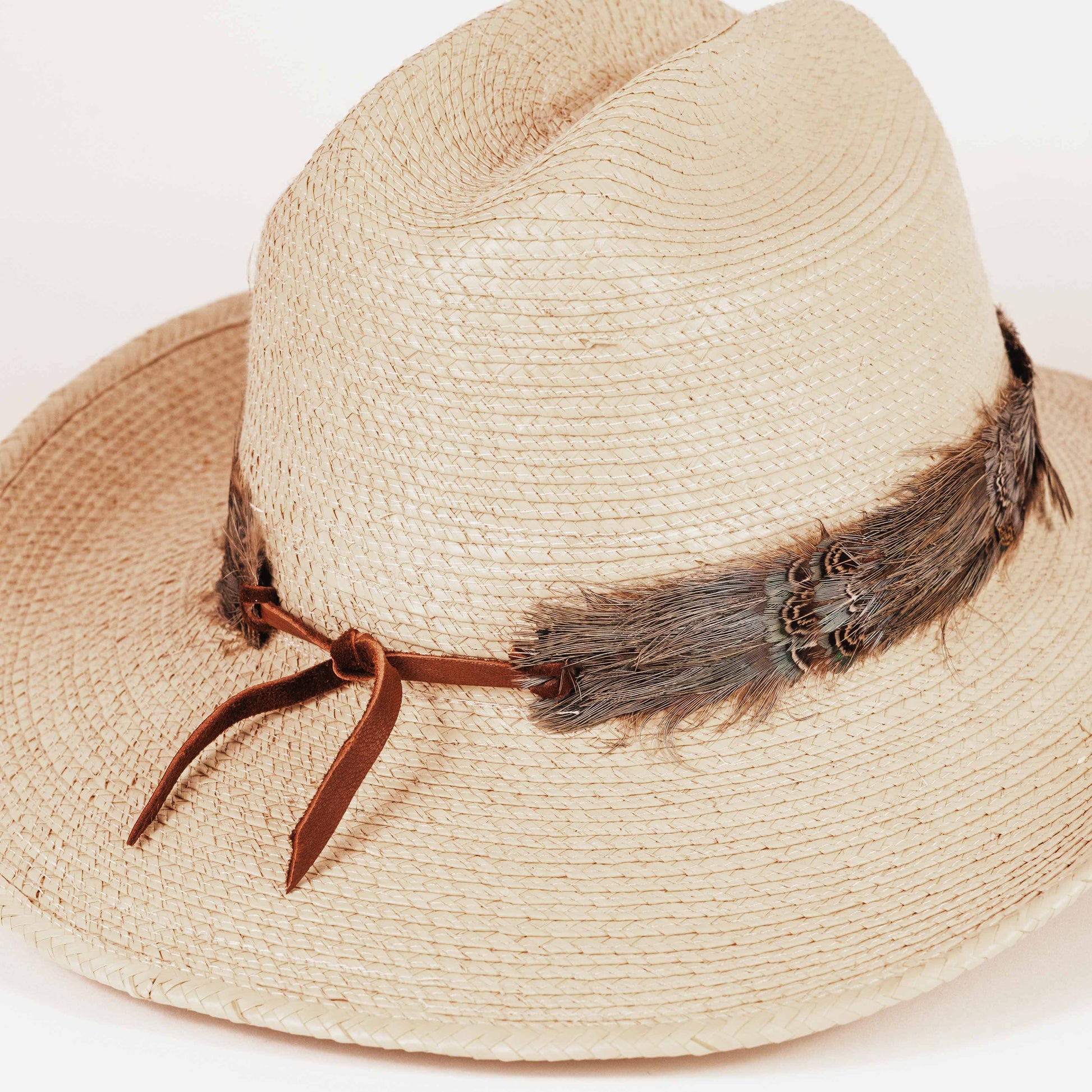 A light beige woven straw hat with a brown leather ribbon band tied in a bow, decorated with dark brown feathers and a small metallic concho accent.