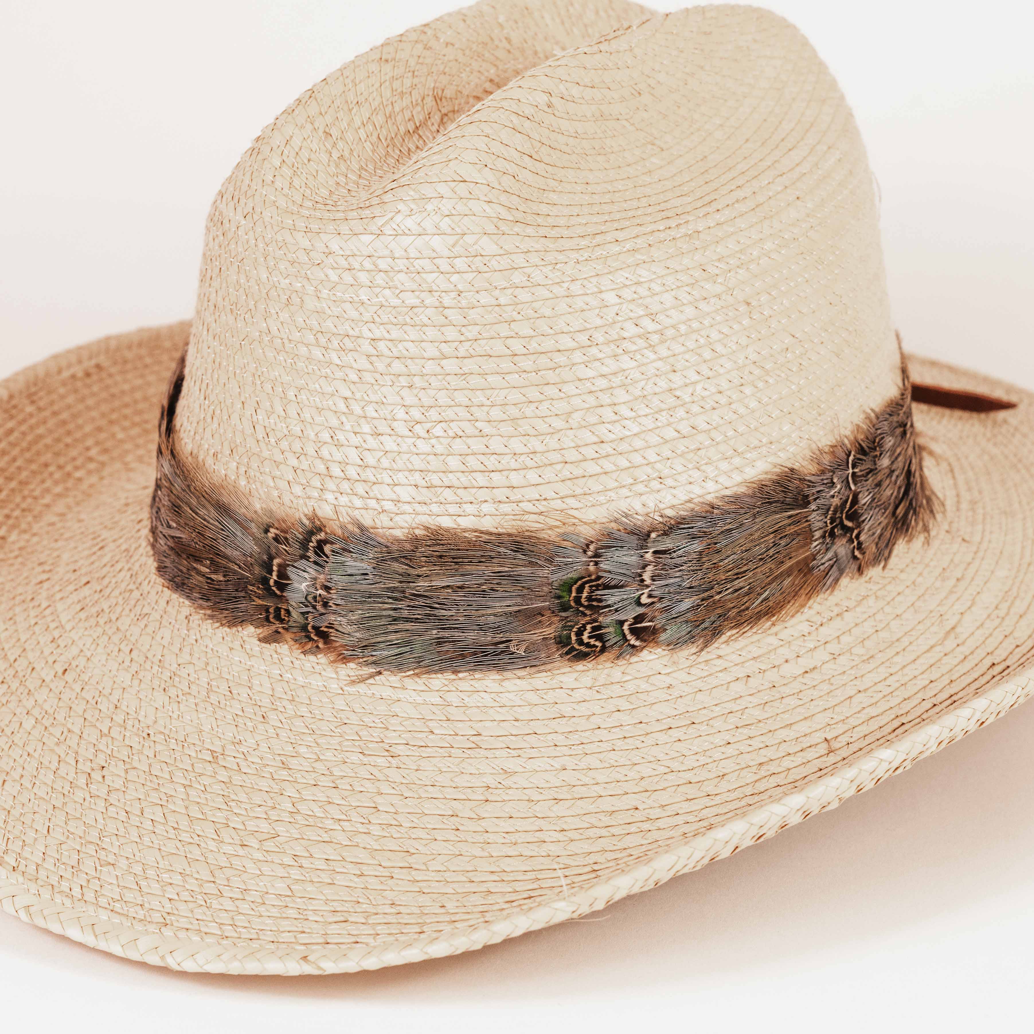 A light beige woven straw fedora hat with a dark brown feather band around the crown, photographed on a clean white background.