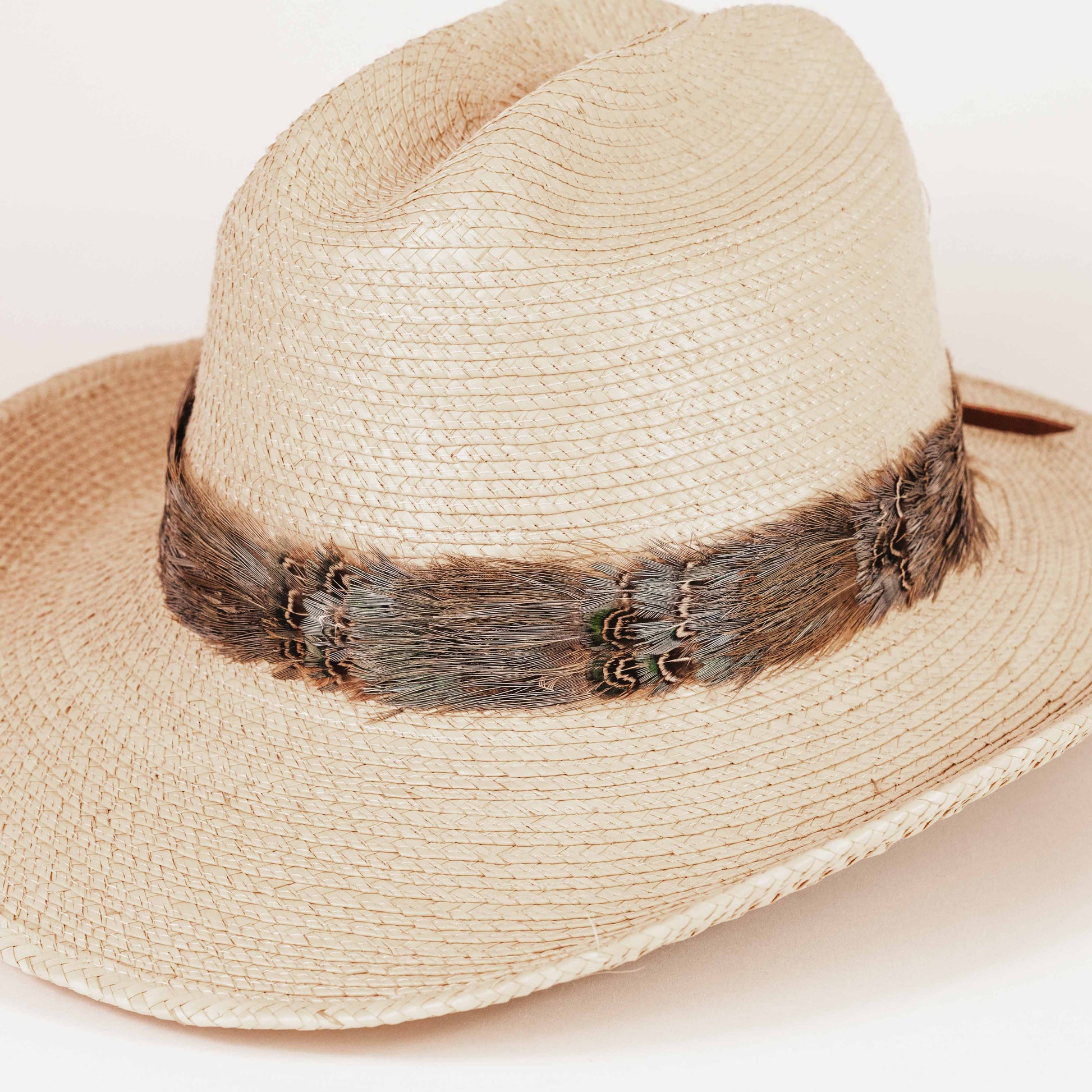 A light beige woven straw fedora hat with a dark brown feather band around the crown, photographed on a clean white background.