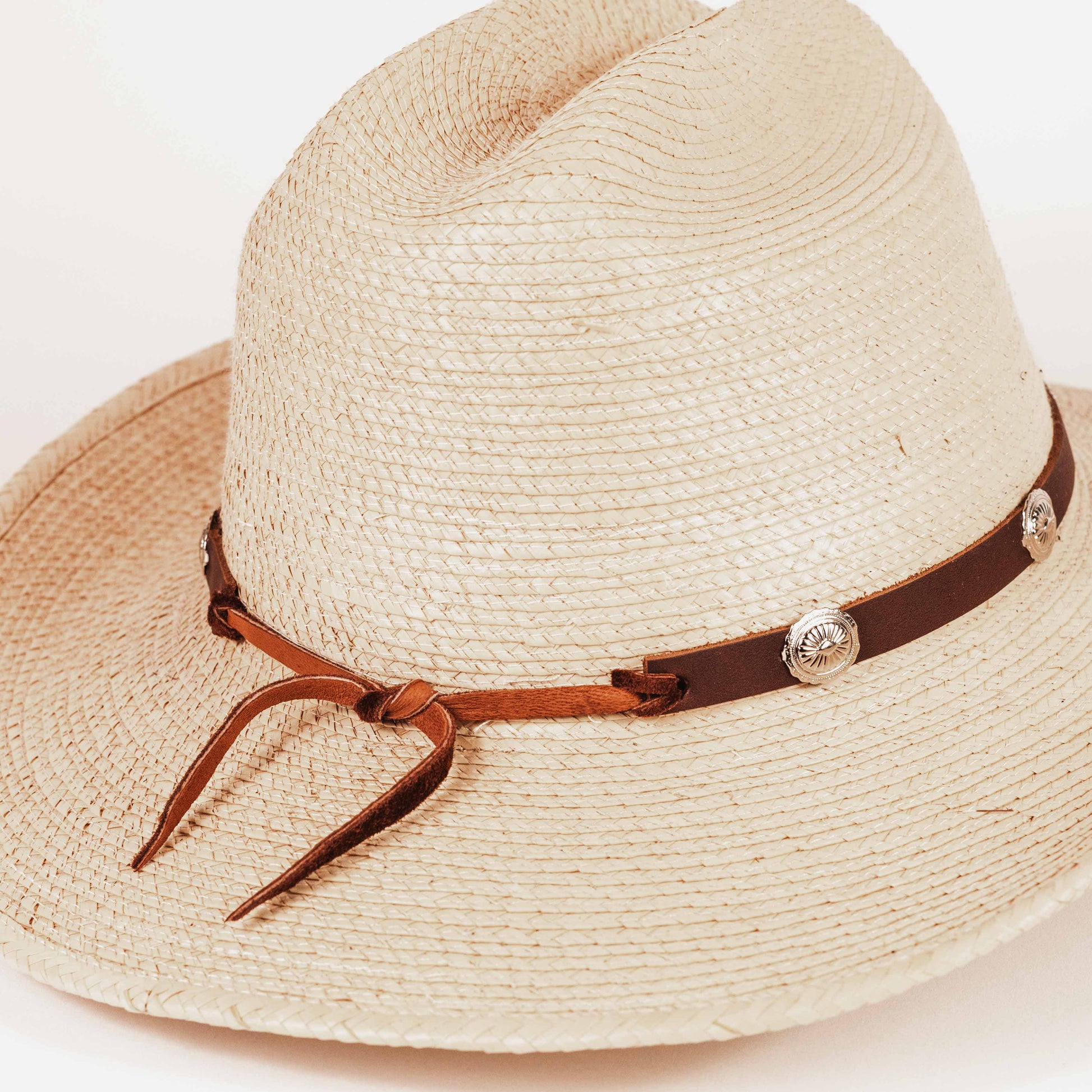 A light beige woven straw western hat with a brown leather band and silver-tone decorative studs, photographed on a clean white background.