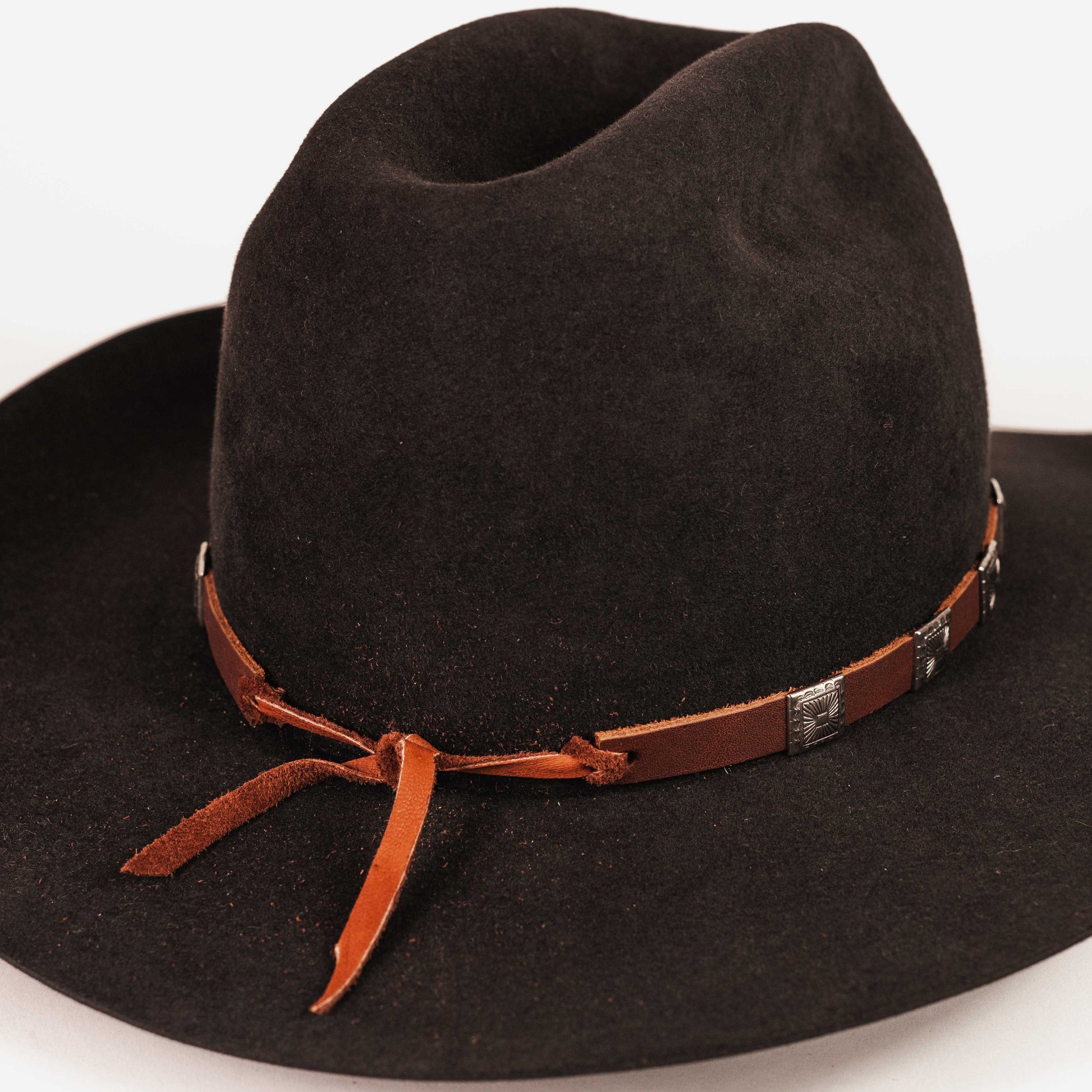 A black felt cowboy hat with a brown leather band tied in a bow and decorated with silver conchos, shown on a plain white background.