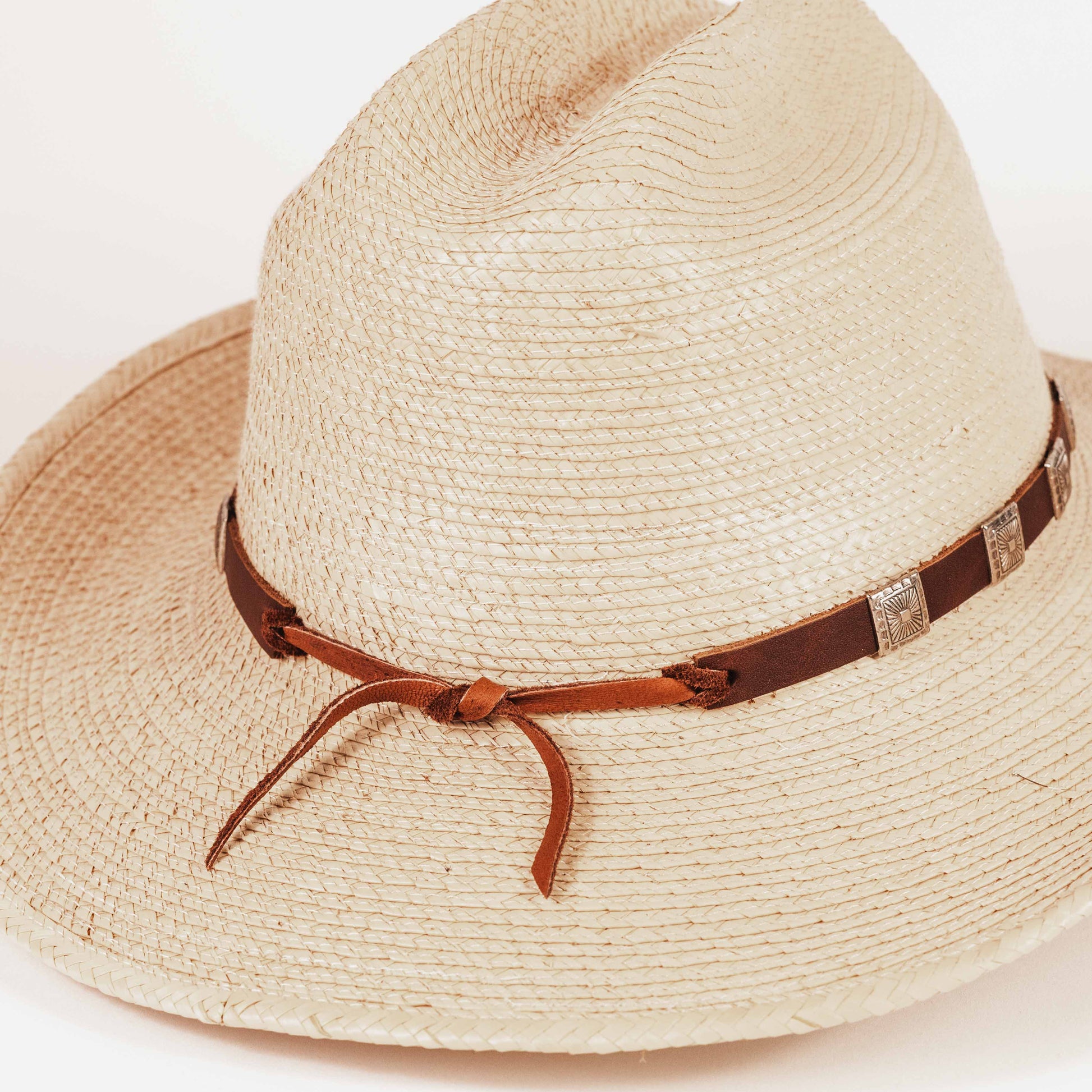 A light beige woven straw cowboy hat with a brown leather band and silver concho accents, photographed on a clean white background.