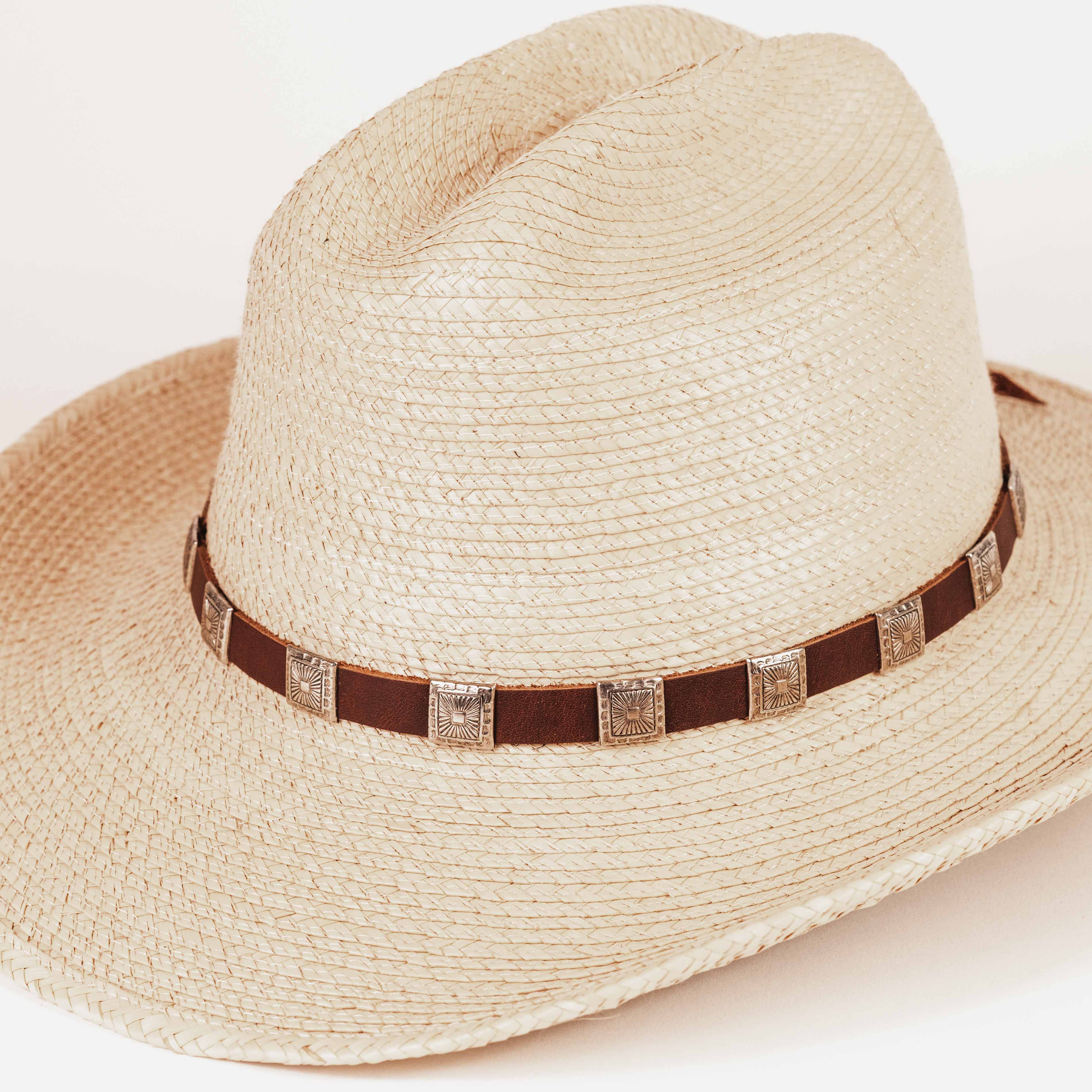 A light beige woven straw western hat with a brown band decorated with square metallic studs, shown on a plain light background.
