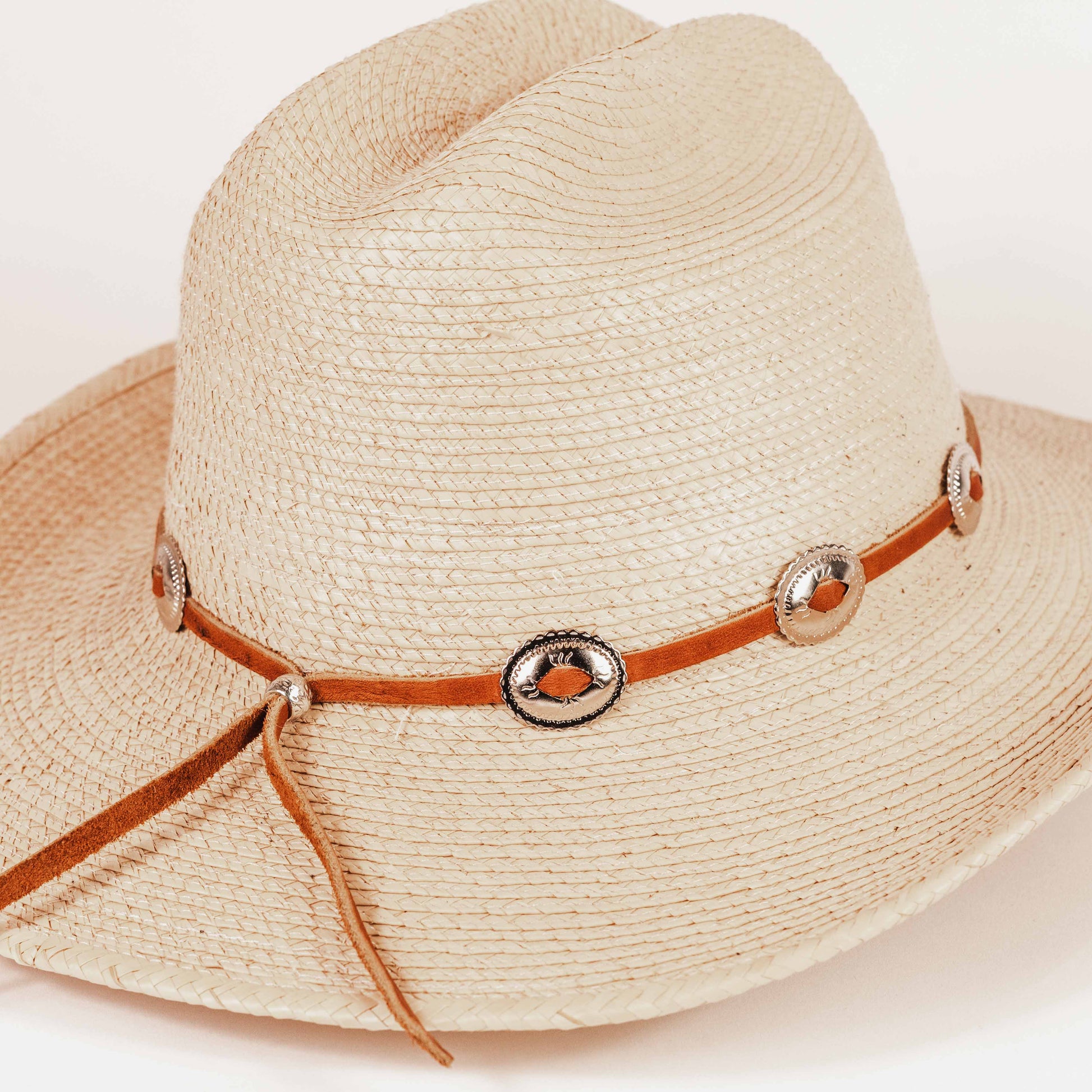 A light beige woven straw cowboy hat with a brown leather band decorated with silver-tone conchos and orange accents, shown on a plain white background.