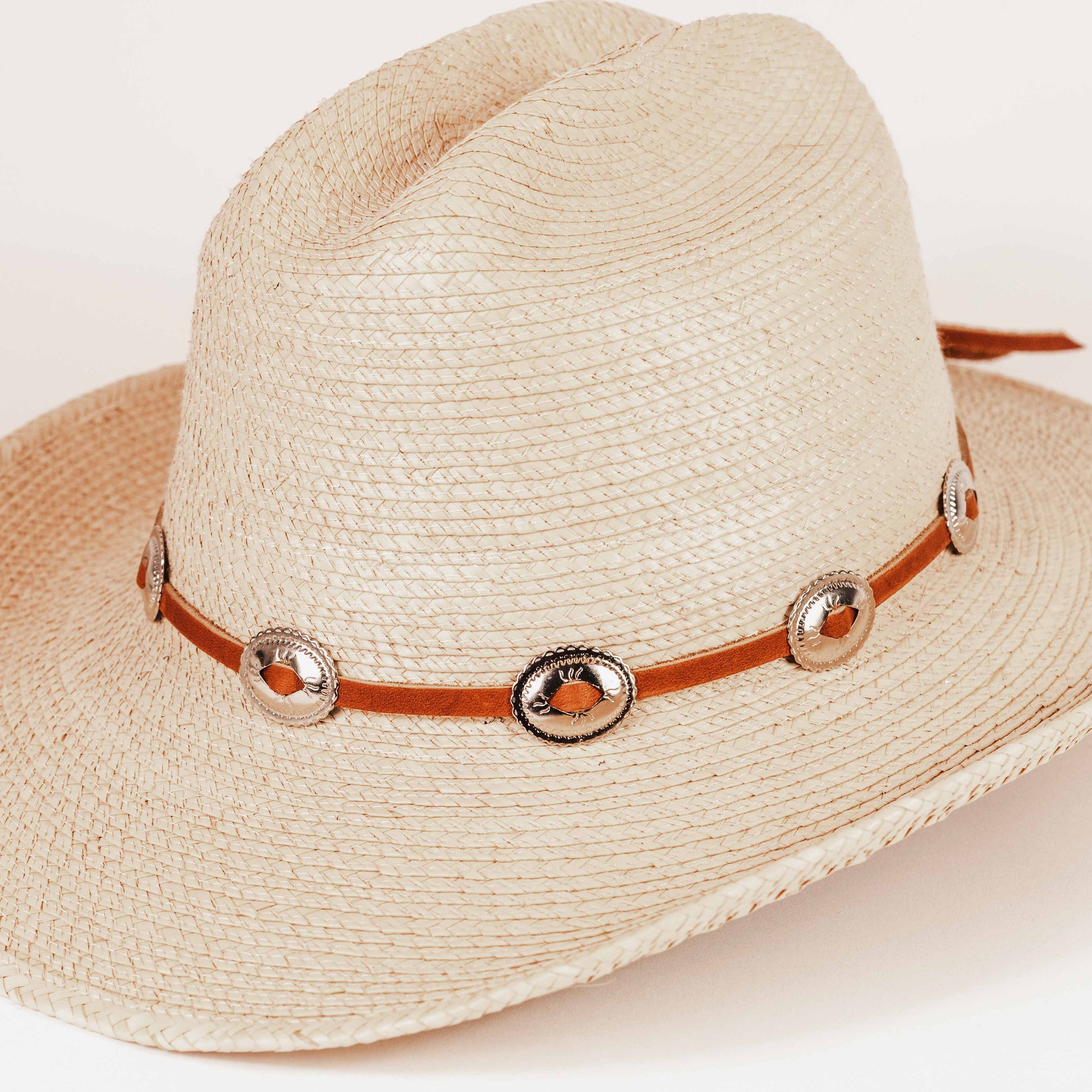 A light beige woven straw cowboy hat with a brown leather band decorated with silver-toned conchos and a small orange accent, shown on a plain light background.