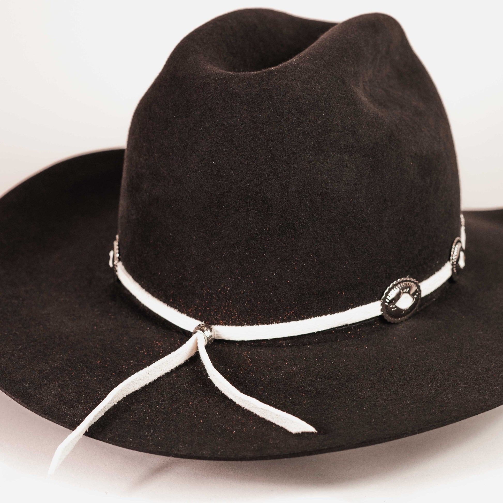A black felt cowboy hat with a white rope band decorated with silver-toned conchos and a white hanging tie, shown on a plain light background.
