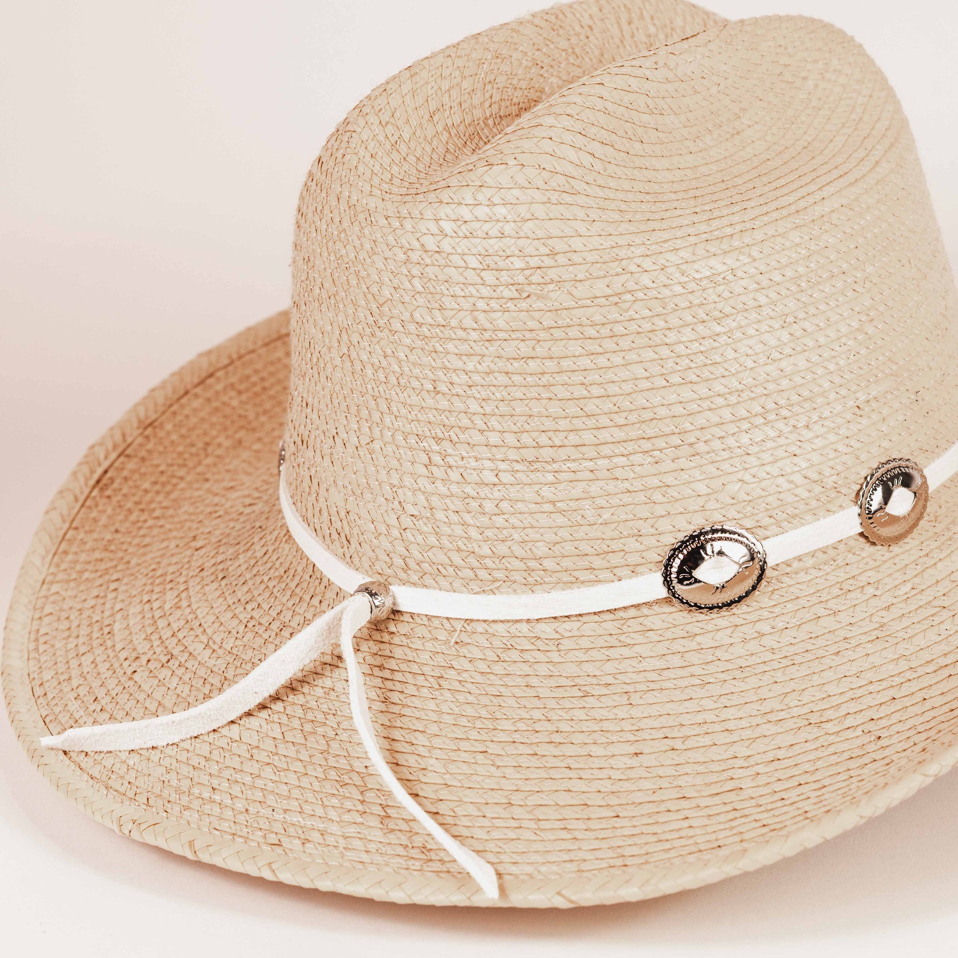 A light beige woven straw hat with a white ribbon band decorated with three metallic silver conchos, shown against a plain light beige background.