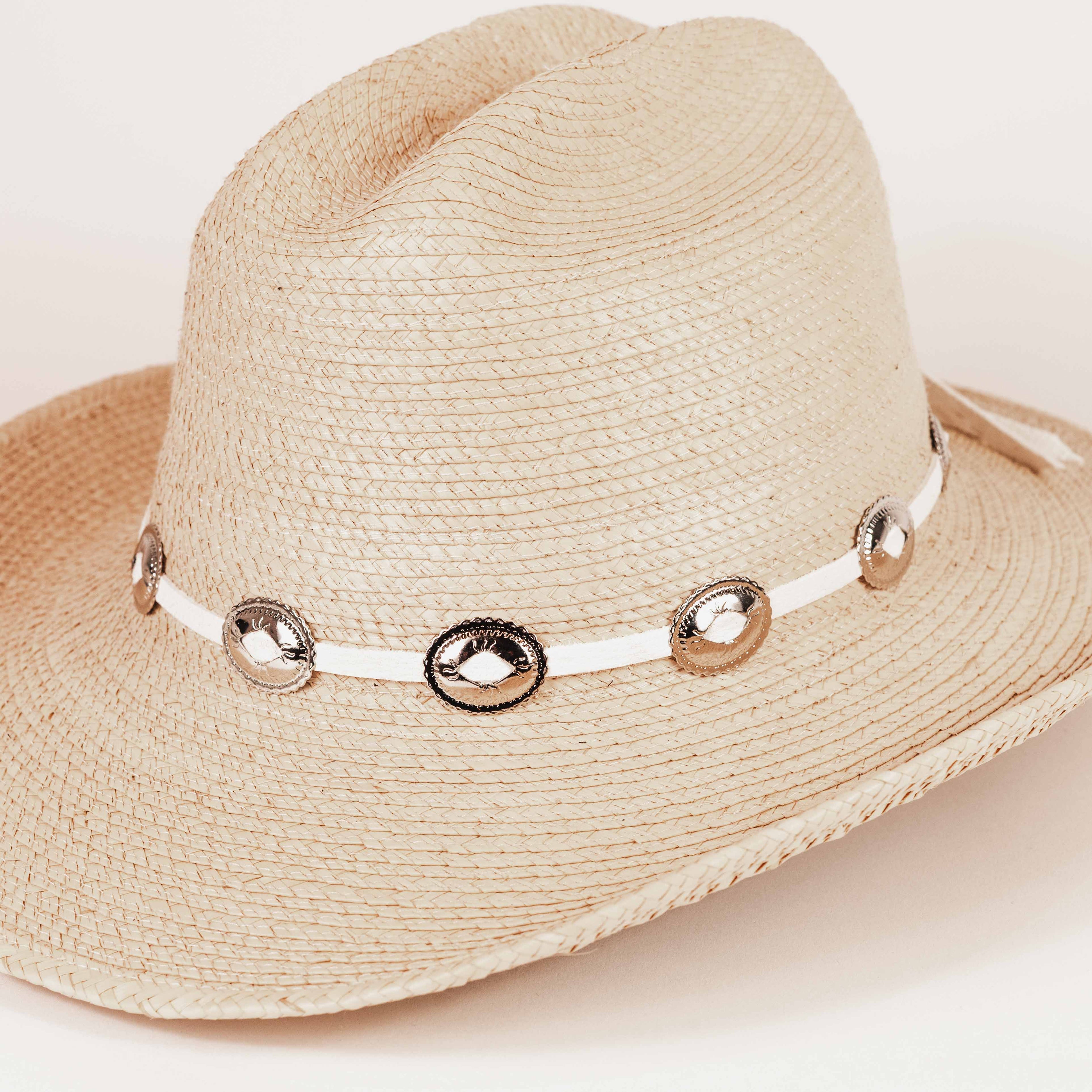 A light beige woven straw hat with a white band decorated with metallic silver conchos, shown on a plain light background.
