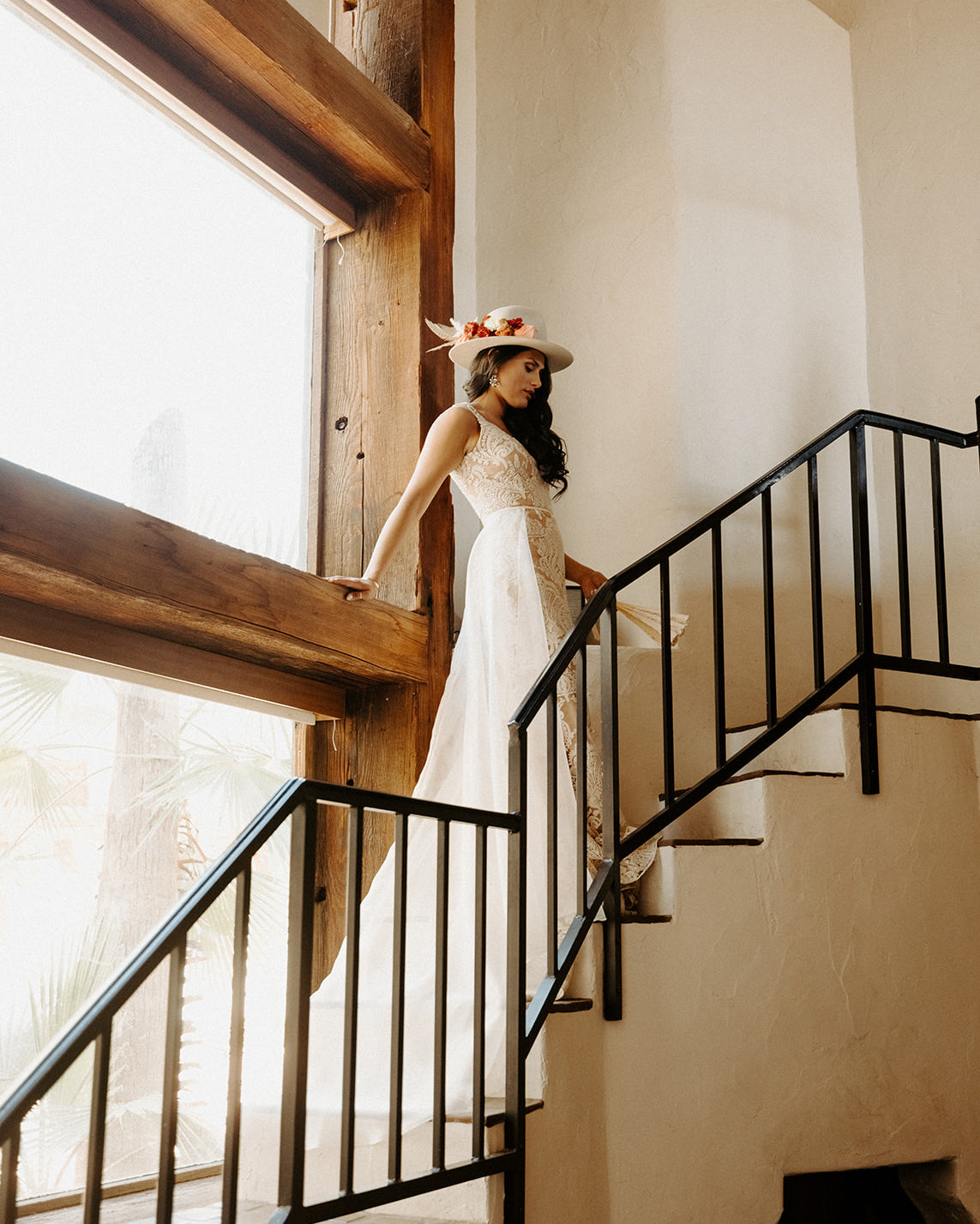 Bride in a wedding dress standing on a staircase with large windows in the background wearing a custom western hat.