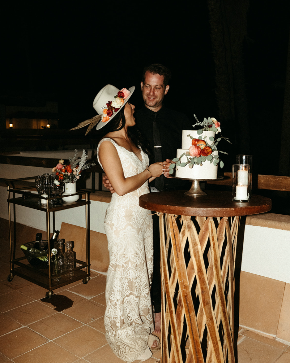 Wedding Couple standing next to a cake on a wooden stand wearing a custom western hatwith a dark background.
