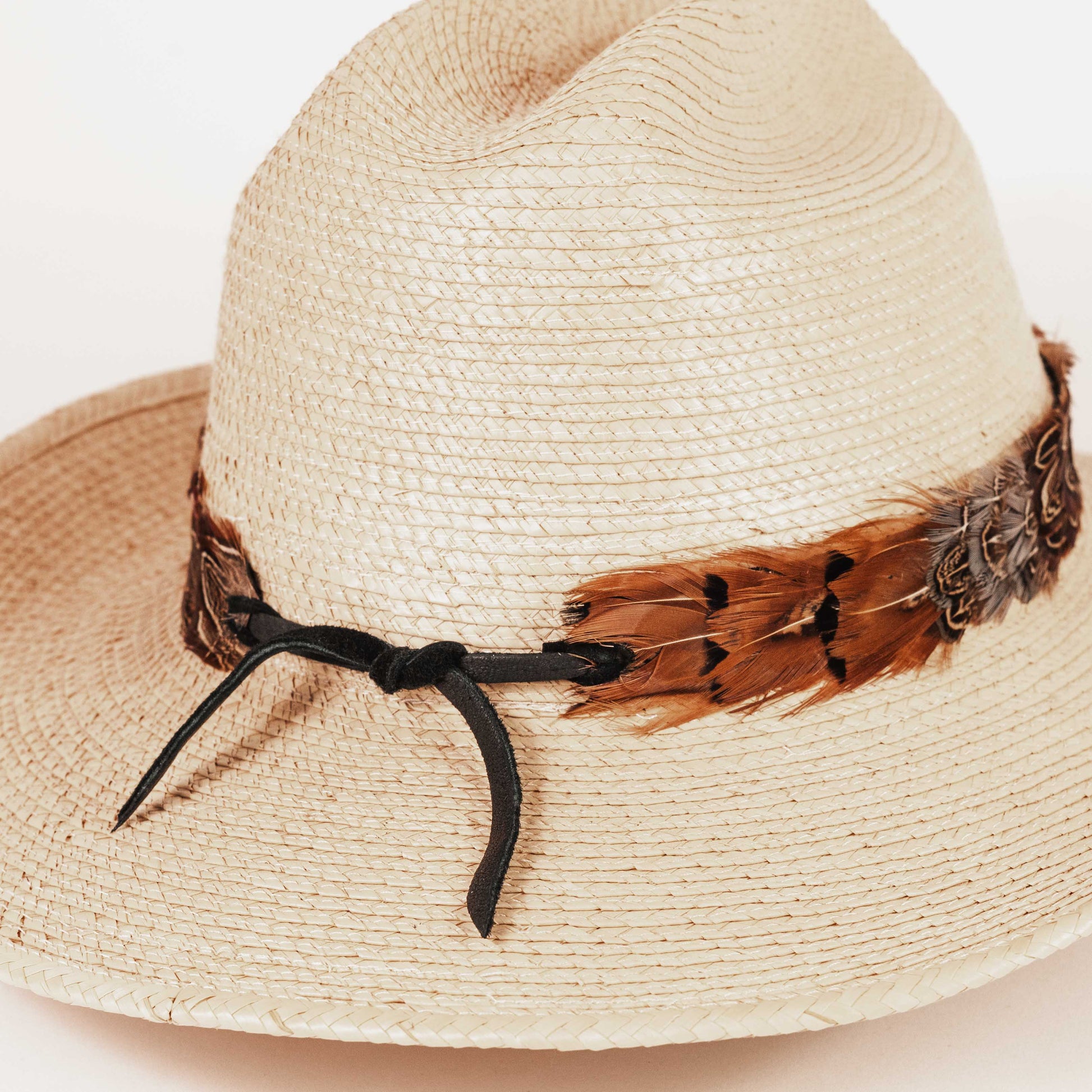A light beige woven straw fedora hat with a decorative brown feather band and a black ribbon tie, shown on a plain light background.