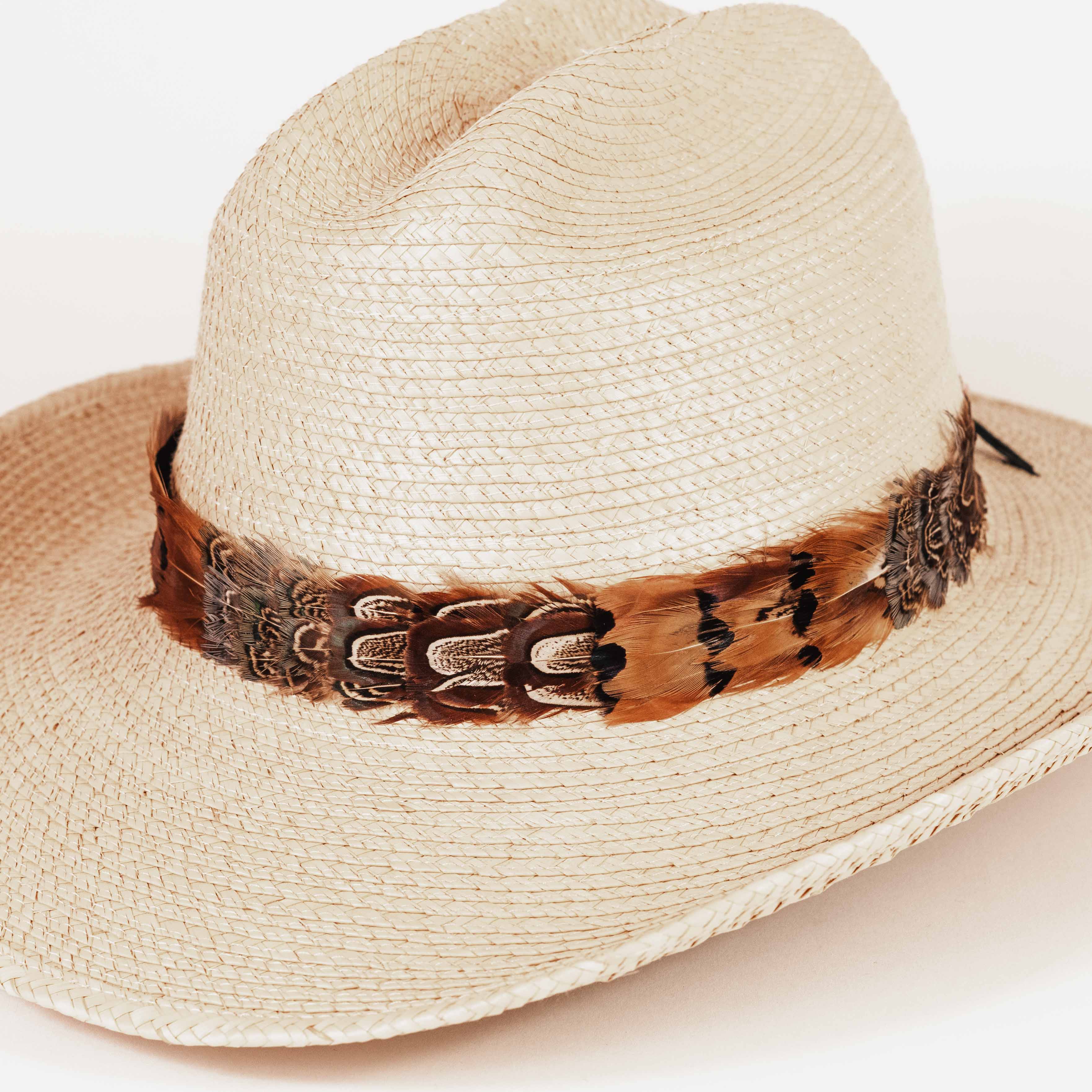 A light beige woven straw cowboy hat featuring a decorative brown and black feather band around the crown, photographed against a clean white background.