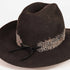 A dark brown felt cowboy hat with a decorative band featuring white and black feather details and a black ribbon tie, shown on a plain white background.
