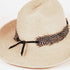 A light beige woven straw hat with a black ribbon bow and a brown-and-white spotted feather band, photographed on a clean white background.