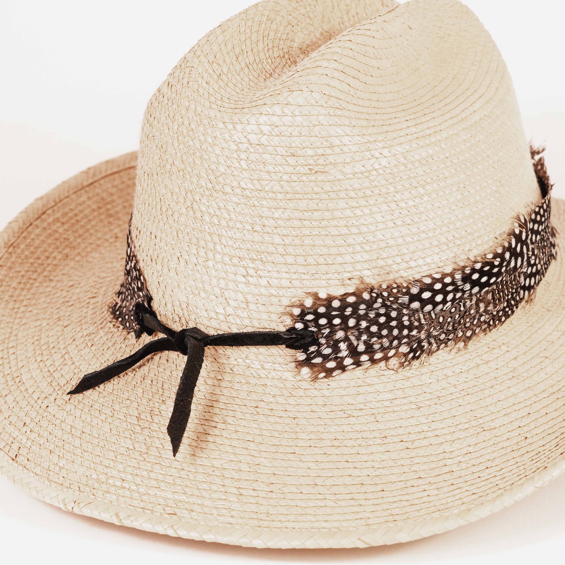 A light beige woven straw hat with a black ribbon bow and a brown-and-white spotted feather band, photographed on a clean white background.