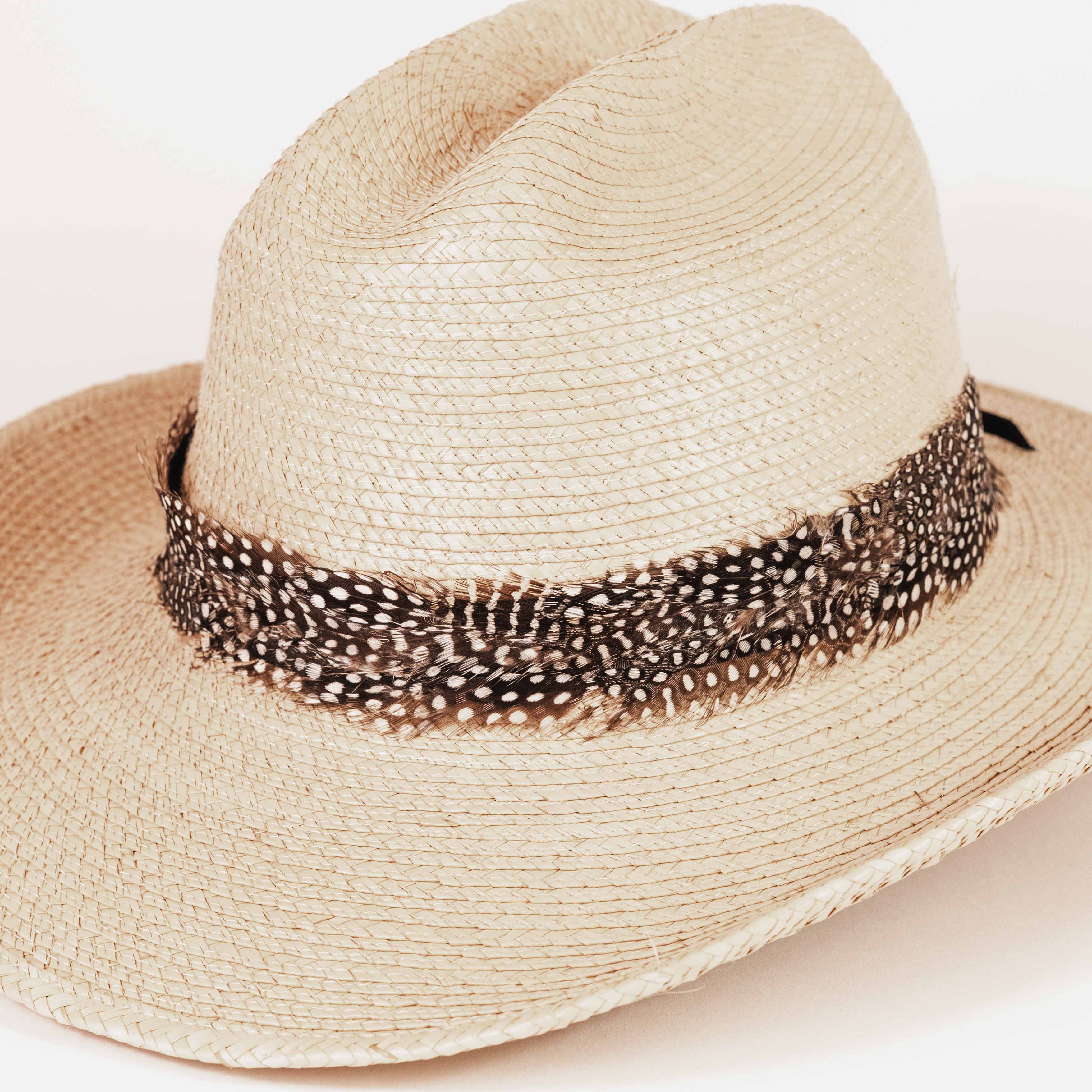 A light beige woven straw western hat with a brown and white spotted feather band around the crown, photographed on a clean white background.