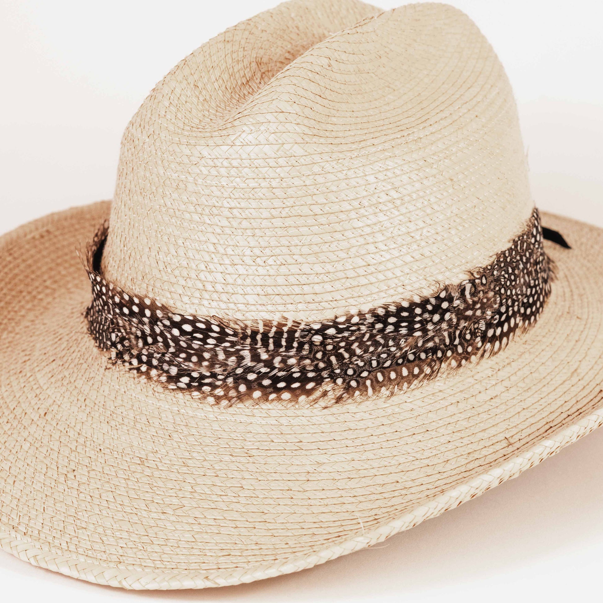 A light beige woven straw western hat with a brown and white spotted feather band around the crown, photographed on a clean white background.