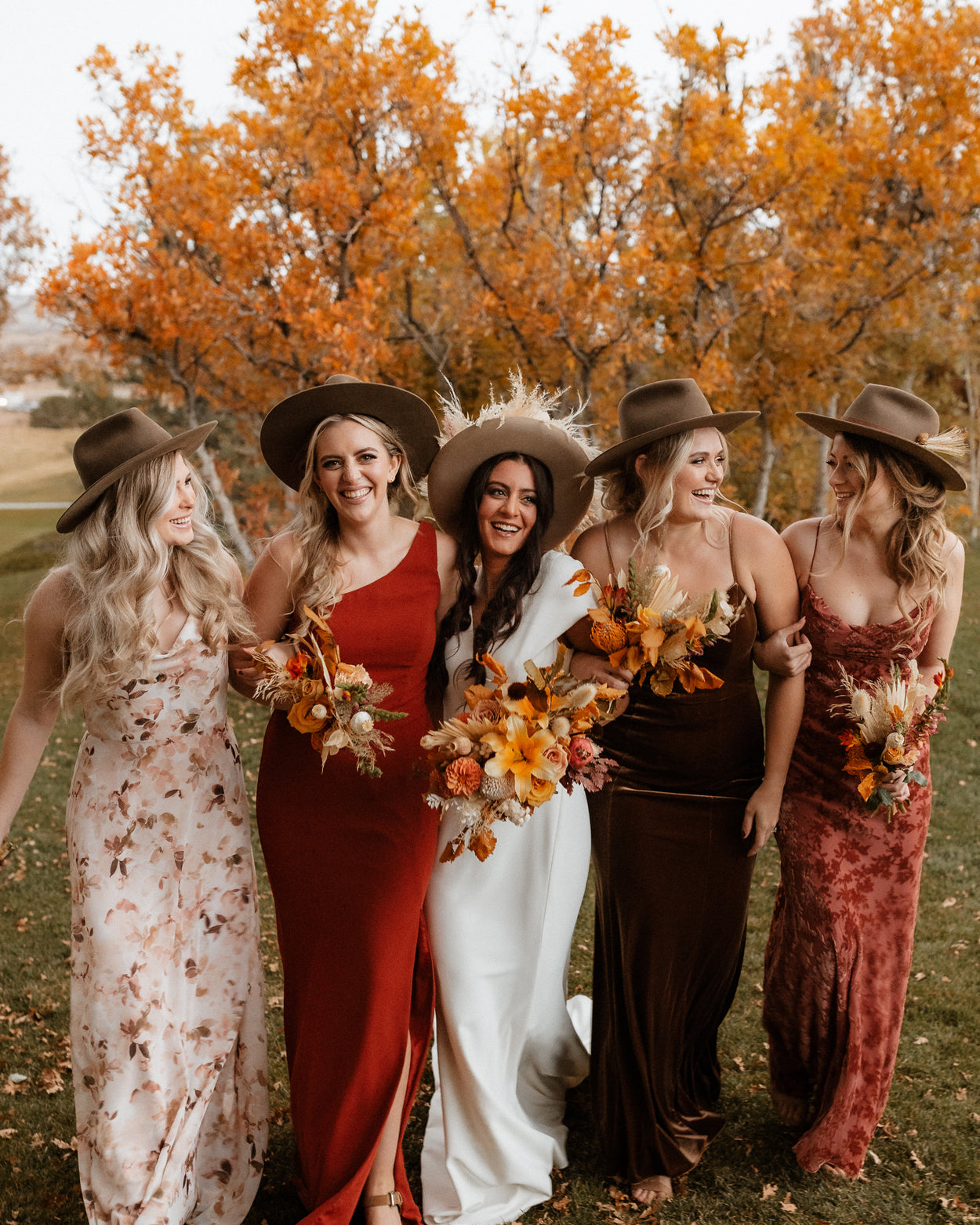 Five bridesmaids in stylish western bjorkstam hats and dresses standing in a field with autumn trees.