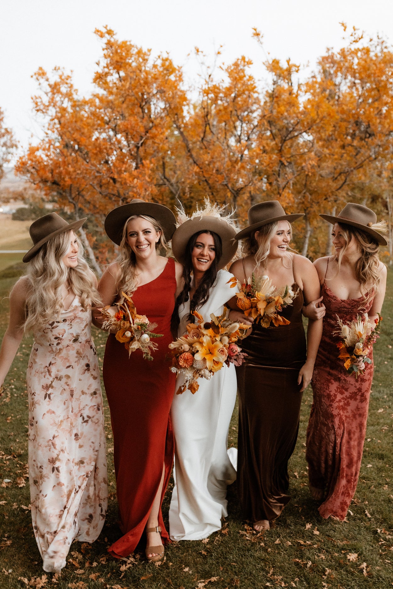 Five bridesmaids in stylish western bjorkstam hats and dresses standing in a field with autumn trees.