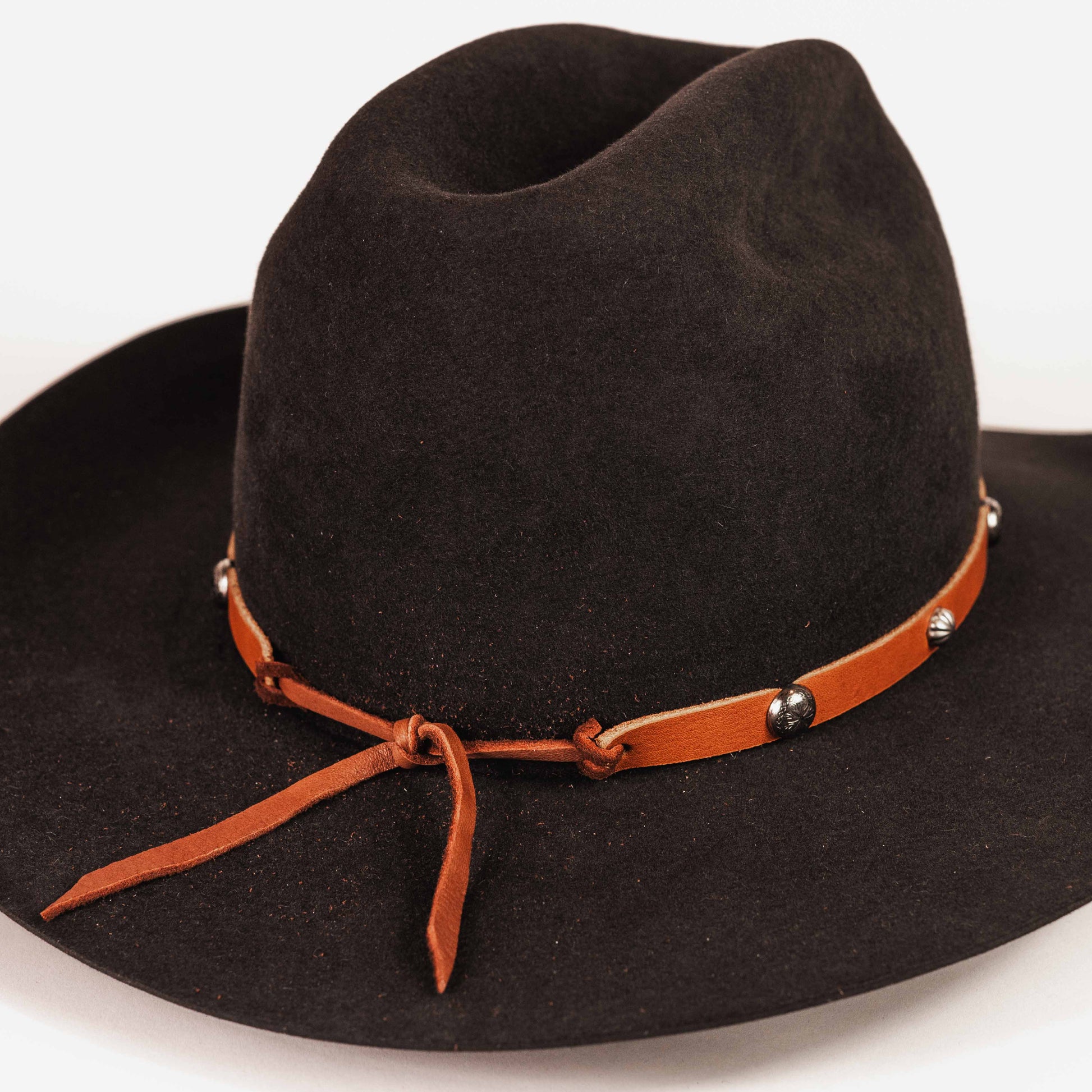 A black felt cowboy hat with a brown leather band tied in a bow and decorated with small silver conchos, shown on a plain white background.