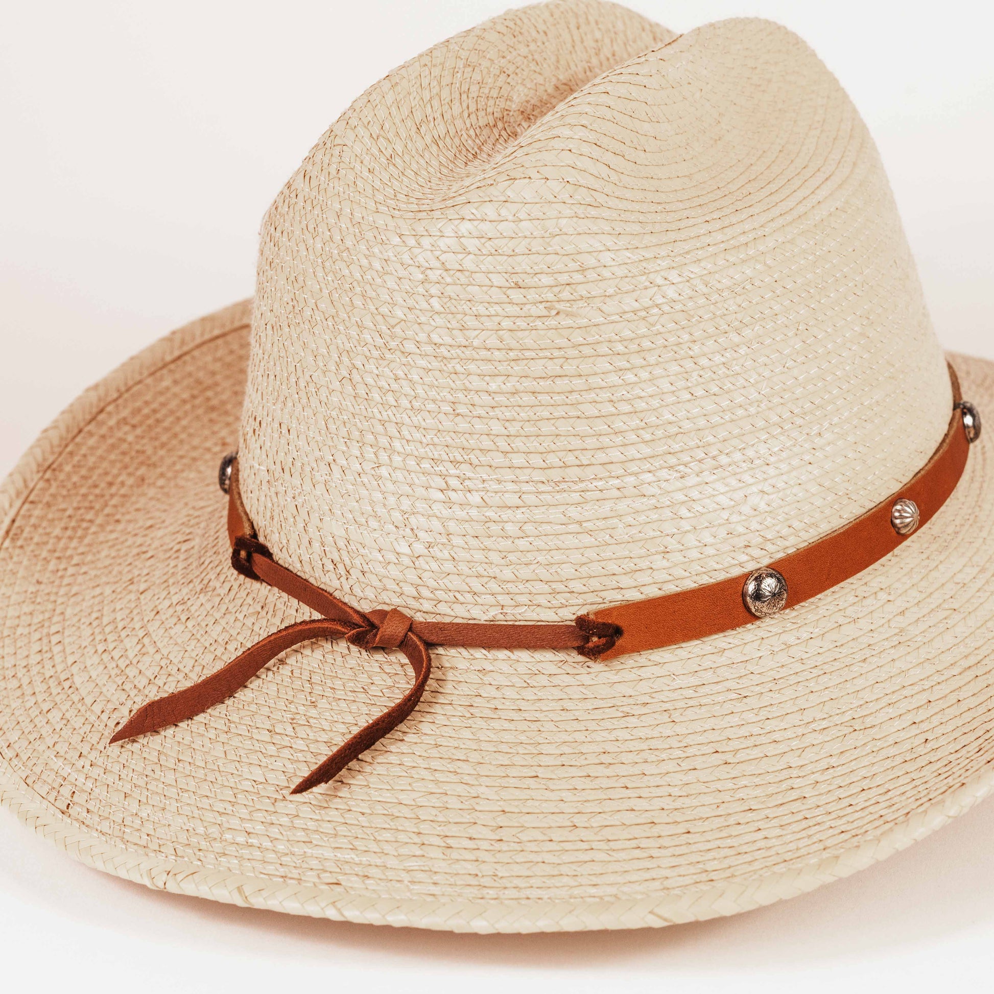 A light beige woven straw fedora hat with a brown leather band decorated with silver-tone studs and a bow, shown on a plain white background.