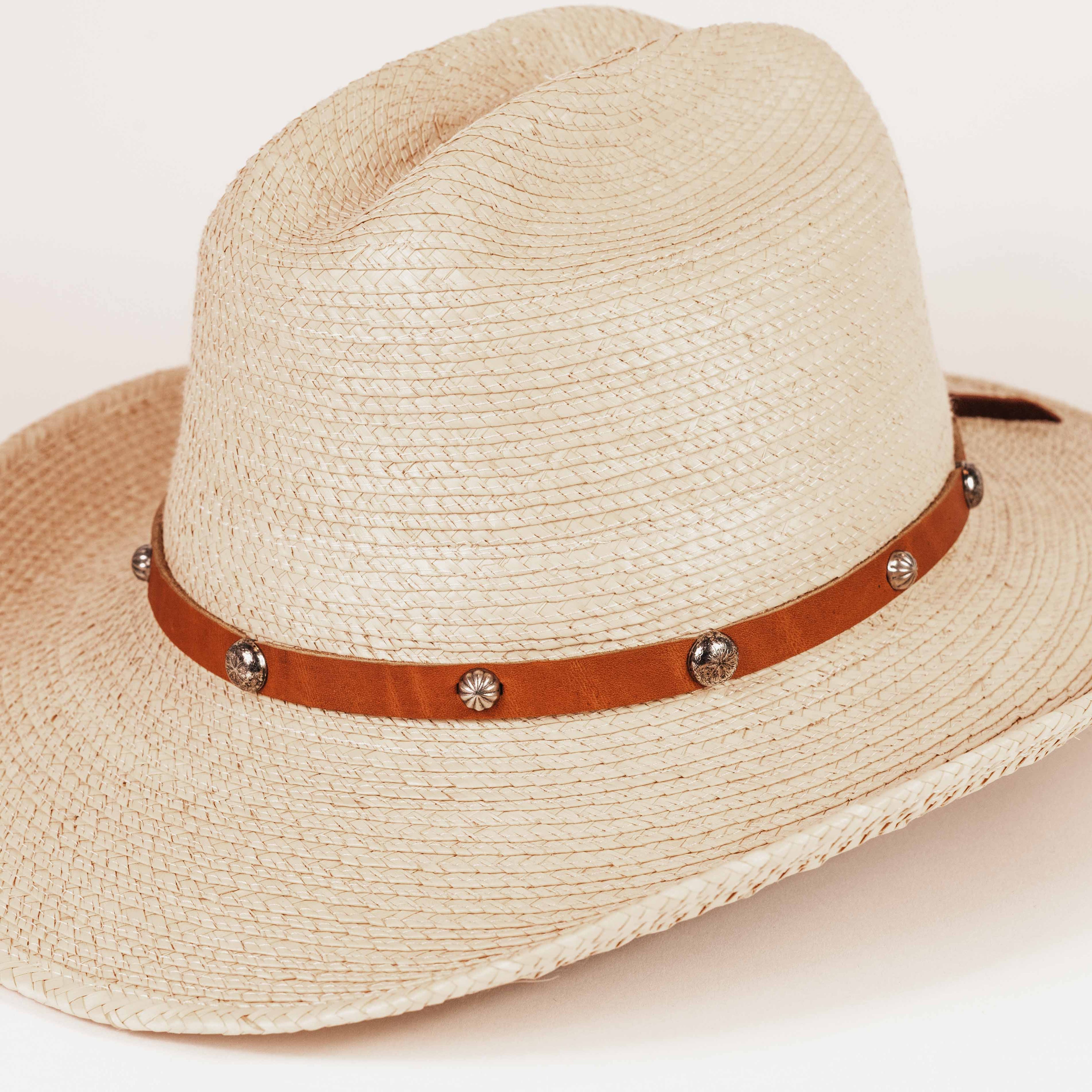 A light beige woven straw cowboy hat with a brown leather band decorated with silver-tone conchos and studs, shown on a clean white background.