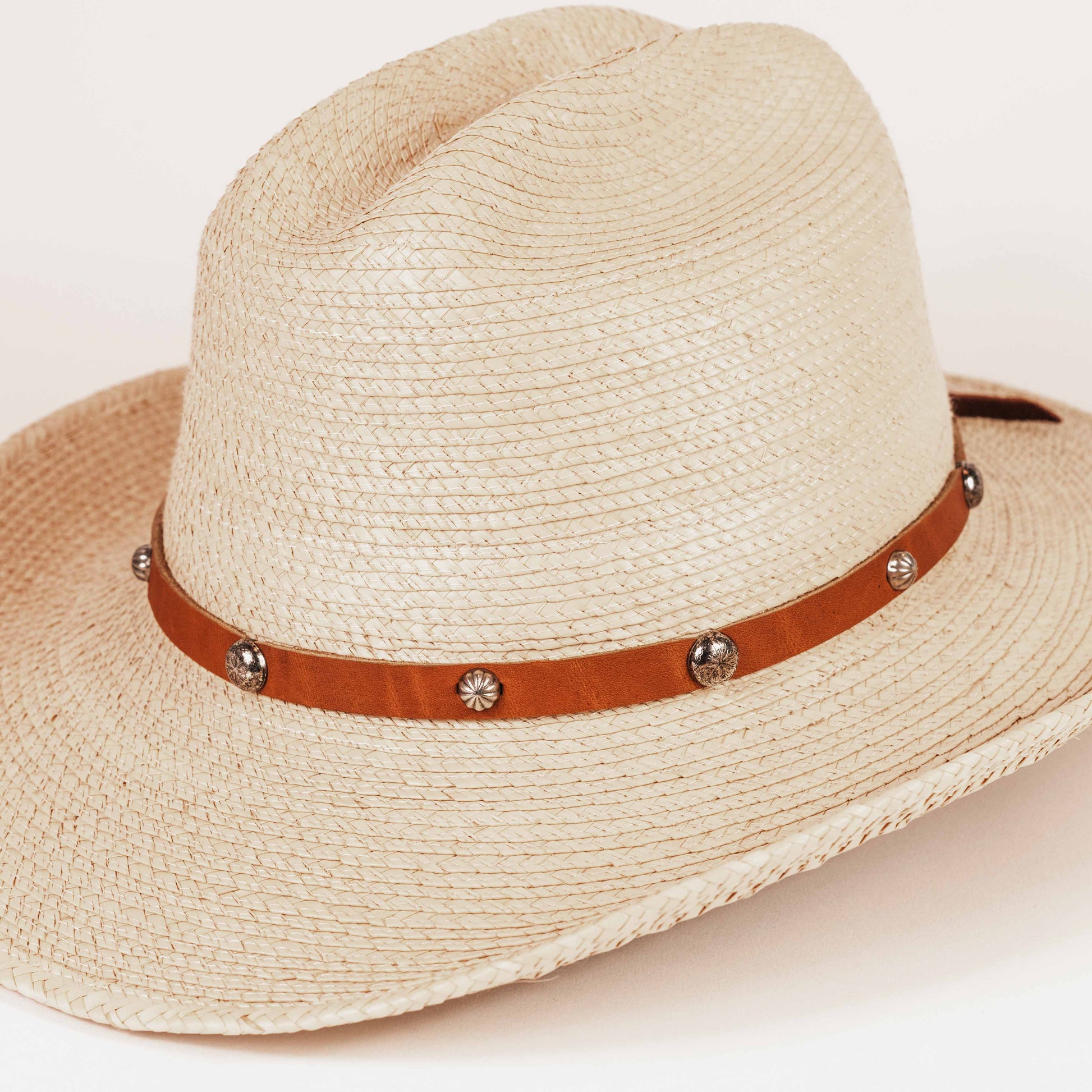 A light beige woven straw cowboy hat with a brown leather band decorated with silver-tone conchos and studs, shown on a clean white background.