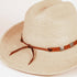 A close-up of a woven straw fedora hat in a light beige color, featuring a brown leather band with silver-toned metal accents and a tied bow, set against a plain white background.
