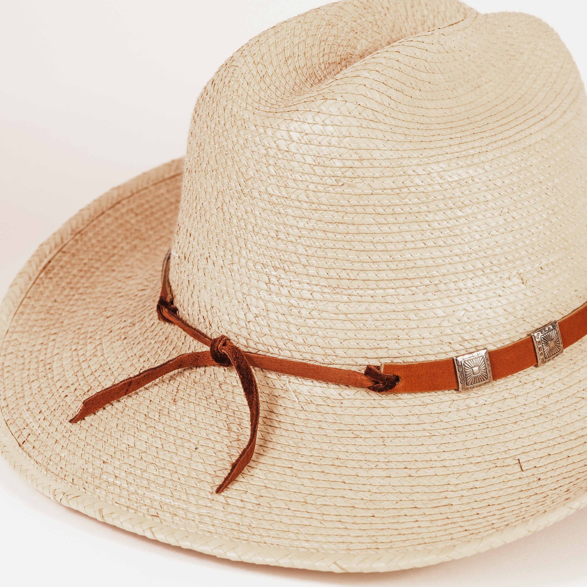 A close-up of a woven straw fedora hat in a light beige color, featuring a brown leather band with silver-toned metal accents and a tied bow, set against a plain white background.