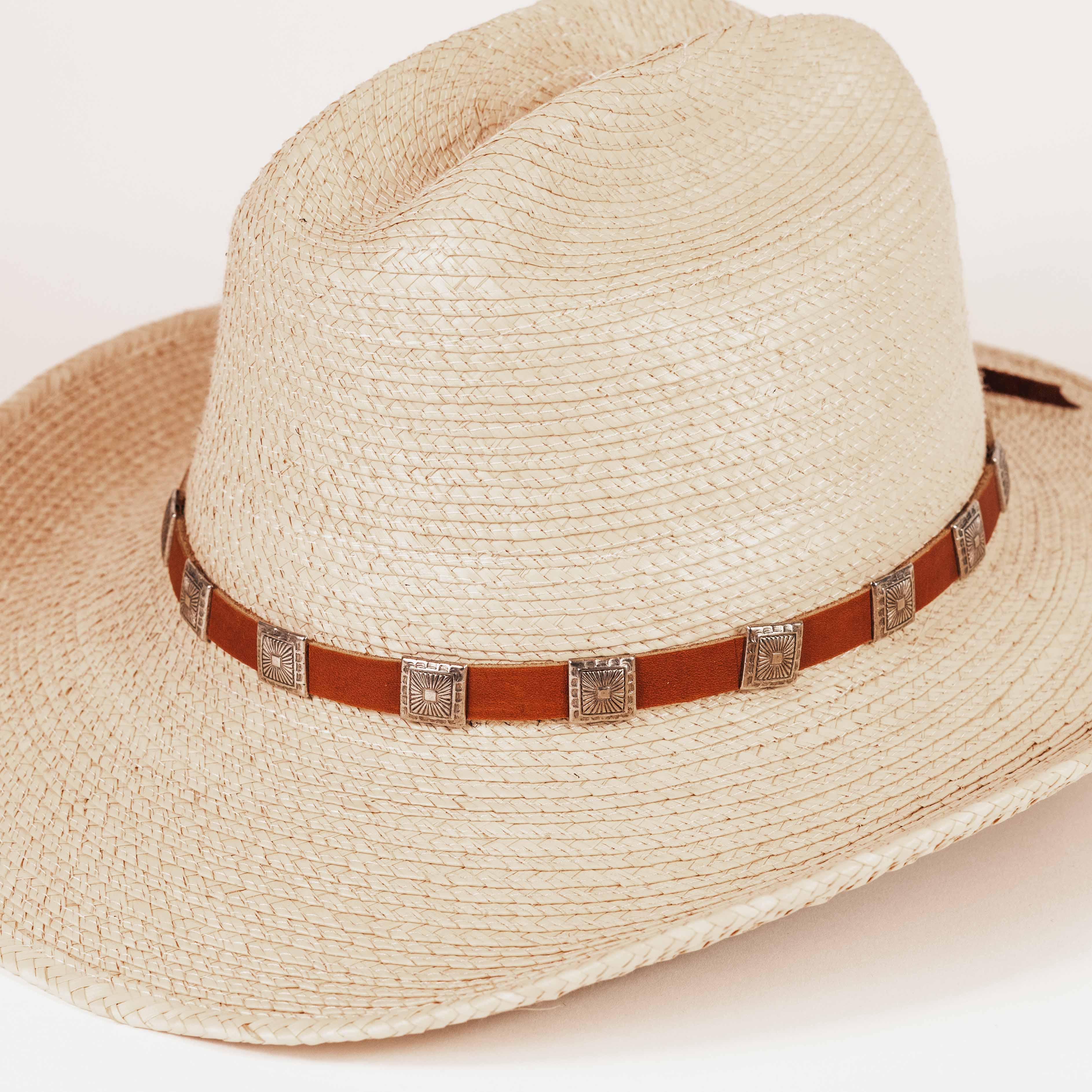 A light beige woven straw cowboy hat with a brown leather band and silver-toned square conchos, photographed on a clean white background.